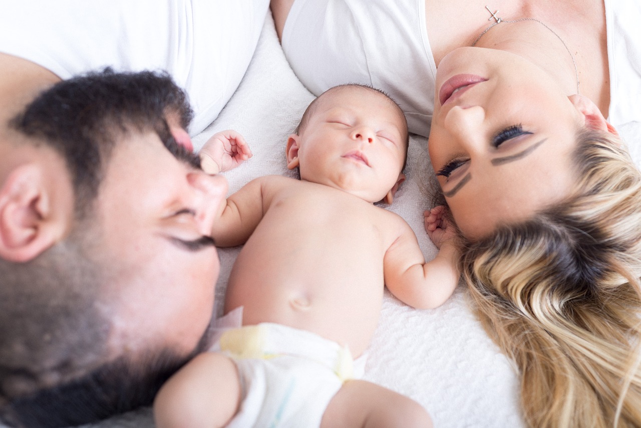 Ein Baby liegt schlafend zwischen einem Mann mit Bart und einer Frau mit blondem Haar. Beide Erwachsene liegen auf der Seite und blicken liebevoll das Baby an