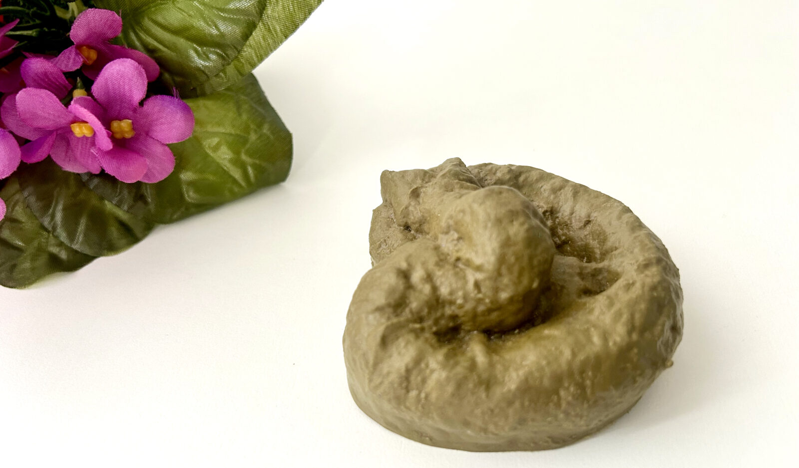 Künstlicher Deko-Gegenstand in Form eines braunen Kothaufens neben einer künstlichen Pflanze mit pinken Blüten.