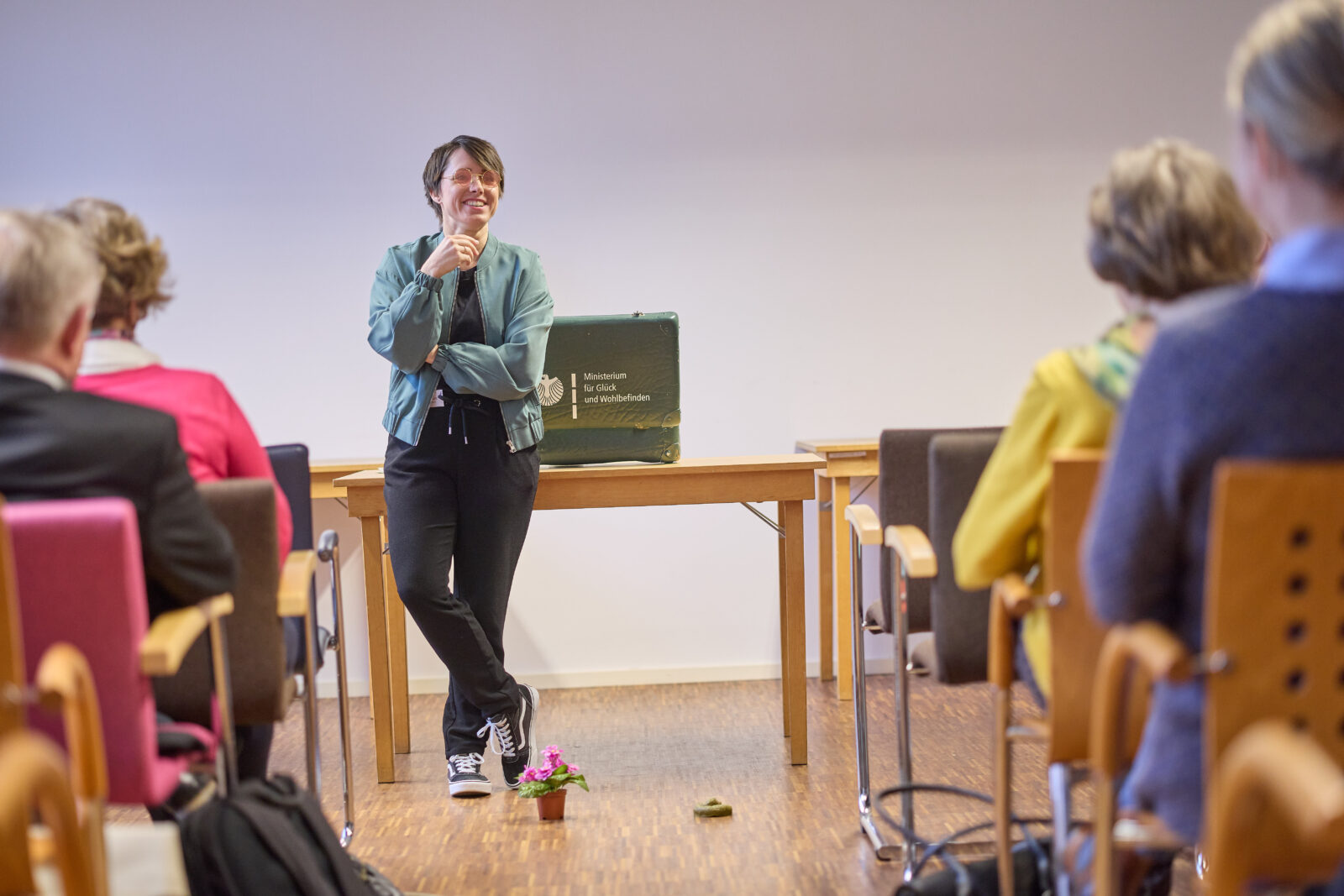 Gina hält einen Vortrag vor einem sitzenden Publikum; im Hintergrund steht ein grüner Koffer mit der Aufschrift ‚Ministerium für Glück und Wohlbefinden‘