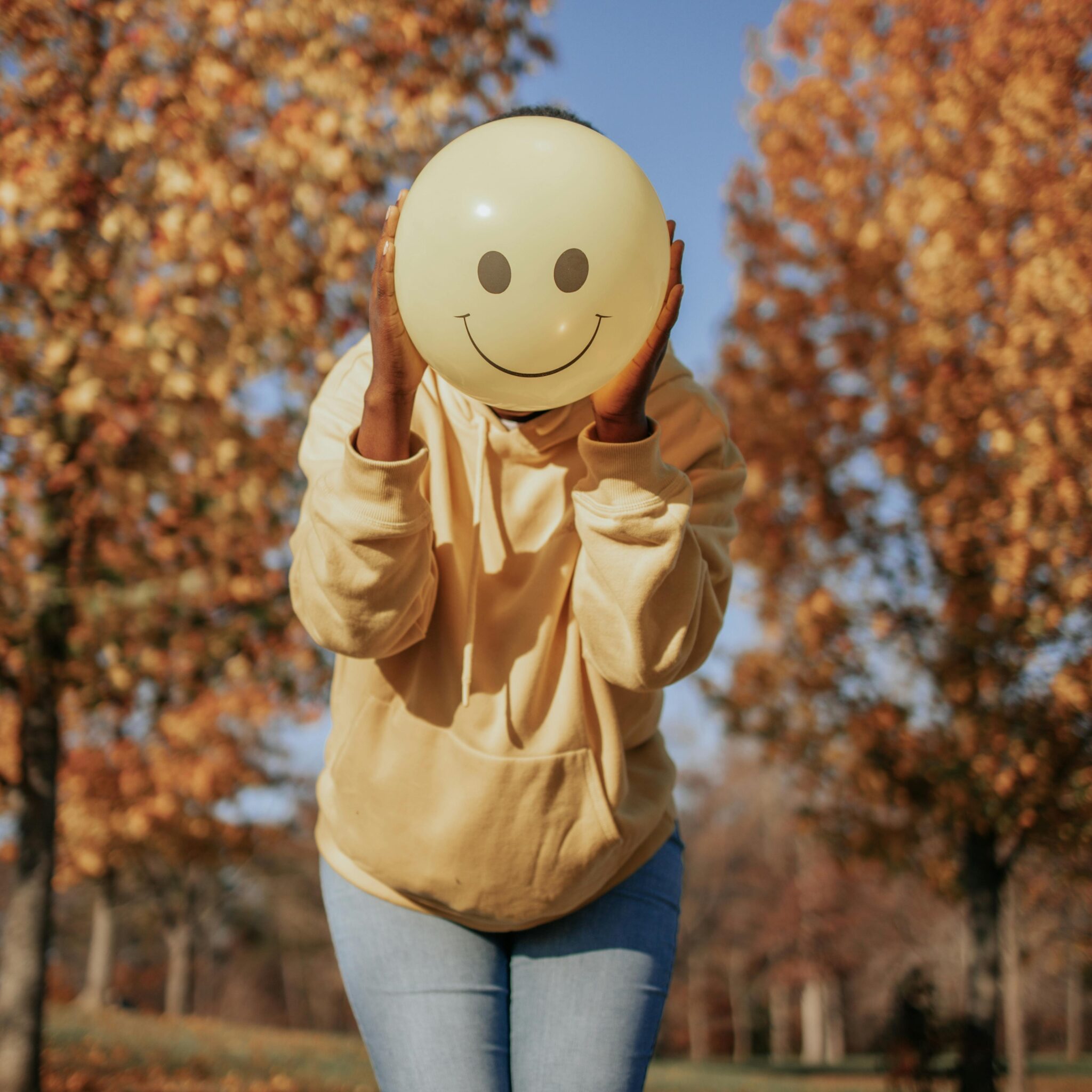 Person hält gelbes Smiley vor dem Gesicht im Park – Symbol für versteckte Gefühle und emotionale Wahrnehmung